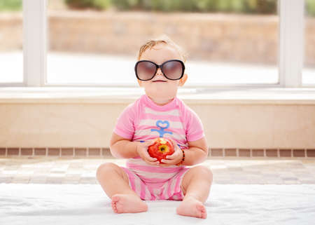Portrait of funny cute white Caucasian smiling baby girl wearing large sunglasses sitting on floor in swimming pool by large window looking in camera, eating appleの写真素材