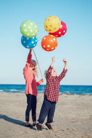 Group portrait of two funny white Caucasian children kids with colorful bunch of  balloons, playing running on beach on sunset, autumn fall seasonの写真素材