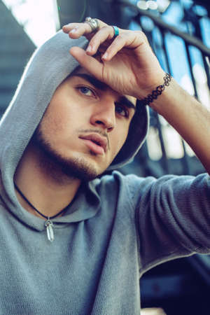 Conceptual art portrait of beautiful handsome gothic young middle east brunette man with blue eyes, beard, wearing blue hoodie, outside in street looking in camera touching his headの写真素材