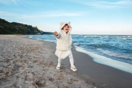 Portrait of one funny smiling laughing white Caucasian child kid baby girl in fur coat and tutu skirt running on ocean sea beach on sunset outdoorsの写真素材