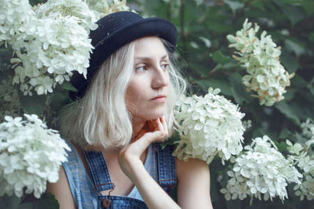 Closeup portrait of beautiful Caucasian teenage young blonde alternative model girl woman in blue tshirt, jeans romper, black hat, sitting among large white flowers on summer dayの写真素材