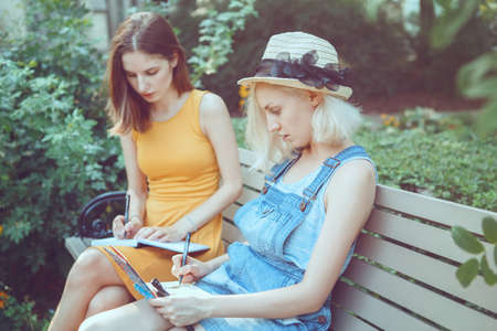 Portrait of two white Caucasian young girls hipster students friends outside in park on summer day sitting on bench together, drawing, sketching, best friends forever, toned with filtersの写真素材