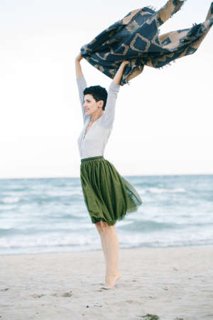 Portrait of beautiful Caucasian white brunette woman with short hair in green olive tutu tulle skirt, with large shawl scarf, standing barefoot on tiptoe on beach looking awayの写真素材