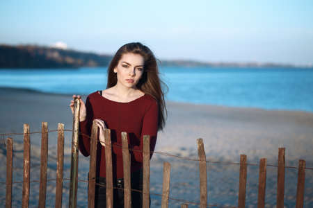 portrait of pensive sad lonely Caucasian young beautiful woman with messy long hair on windy autumn fall day outdoor on the shore beach near wooden fence at sunset, looking in cameraの写真素材