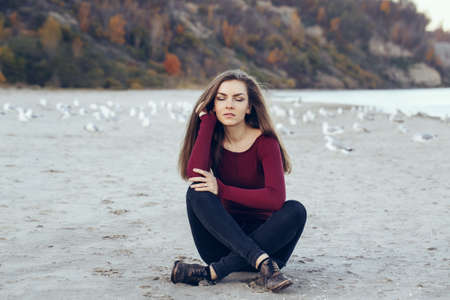 portrait of  Caucasian young beautiful woman with closed eyes, long hair, wearing black jeans and red shirt, sitting on sand on beach among seagulls birds on autumn fall day outdoor at sunsetの写真素材