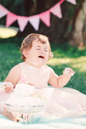 Portrait of cute funny upset sad crying Caucasian baby girl in pink tutu dress celebrating her first birthday with gourmet cake outside in parkの写真素材
