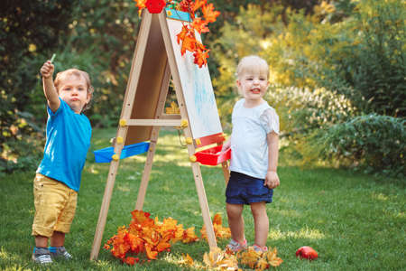 Group of two white Caucasian toddler children kids boy and girl standing outside in summer autumn park by drawing easel with markersの写真素材