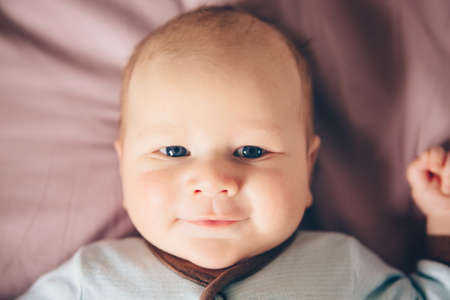 Closeup portrait of cute adorable funny white Caucasian blond little baby boy newborn with blue grey eyes lying on bed looking in camera smiling, lifestyle candid, real lifeの写真素材