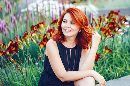 Closeup portrait of smiling middle aged white caucasian woman with waved curly red hair in black dress looking away outside in park garden among flowersの写真素材
