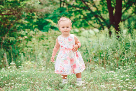 Cute adorable white Caucasian baby girl child in white dress standing in green summer park forest outside,looking in cameraの写真素材