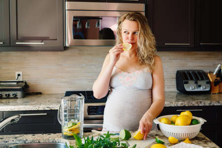 Portrait of white Caucasian blonde pregnant woman eating citrus lemon making juice standing in kitchen at home looking away, healthy lifestyle concept, pregnancy cravingsの写真素材