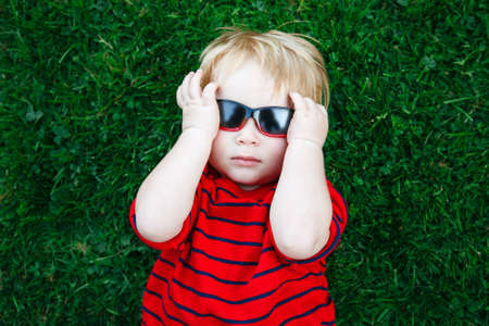 Close up portrait of funny cute adorable white Caucasian toddler child boy with blond hair in red pullover sunglasses lying on green grass. View from above top overhead.の写真素材
