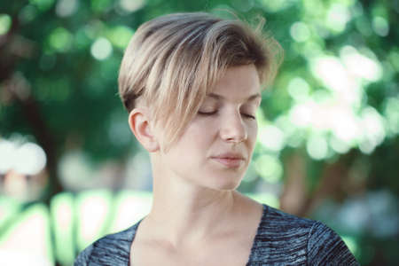 Closeup portrait of calm sad young middle aged white caucasian girl woman with short hair stylish haircut in t-shirt with closed eyes outside in summer parkの写真素材