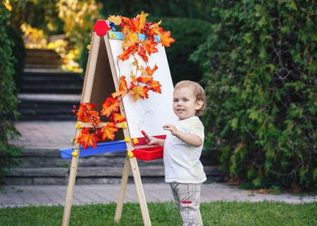 Portrait of white Caucasian toddler child kid boy standing outside in summer autumn park drawing on easel with markersの写真素材