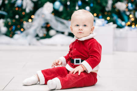Portrait of blond Caucasian baby boy child with blue eyes in red Santa Claus costume sitting by New Year tree, lifestyle Christmas holiday conceptの写真素材