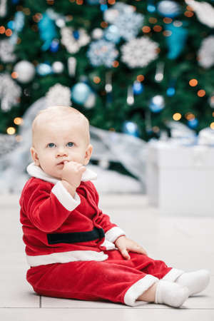Portrait of blond Caucasian baby boy child with blue eyes in red Santa Claus costume sitting by New Year tree, lifestyle Christmas holiday conceptの写真素材
