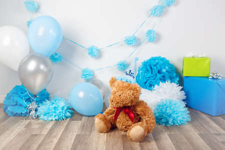 Festive background decoration for first year birthday celebration with little bear toy sitting on floor in studio, balloons, paper flowers and presents gift boxesの写真素材