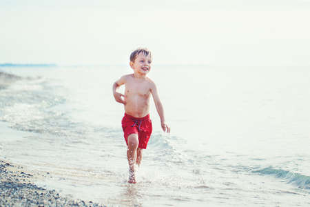 Portrait of funny adorable white Caucasian one young little boy in red swim shorts running on beach by water ocean sea, emotional healthy active lifestyle summerの写真素材