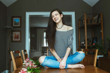 Portrait of smiling laughing Caucasian young beautiful woman model with messy long hair in ripped blue jeans and striped t-shirt sitting on table with flowers indoor looking away, toned with filtersの写真素材