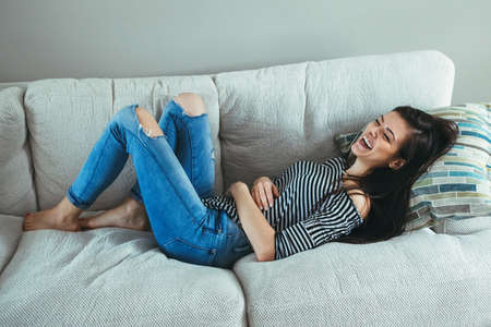 Portrait of smiling laughing Caucasian young beautiful woman model with messy long hair in ripped blue jeans and striped t-shirt lying on sofa couch at home indoor, real lifestyle candidの写真素材