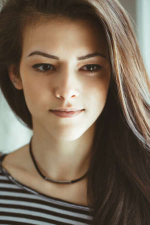 Closeup portrait of smiling Caucasian brunette young beautiful woman model with long hair and brown eyes in striped t-shirt  by window indoor, toned with filtersの写真素材