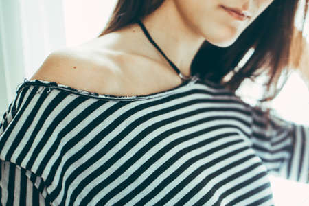 Closeup portrait of Caucasian young beautiful woman model with messy long hair in striped t-shirt by window indoor showing one bare shoulder, toned with filtersの写真素材
