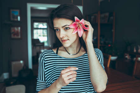 Closeup portrait of smiling pensive Caucasian young beautiful woman model with long hair in striped t-shirt holding large pink flower near ear hair, looking away, toned with filtersの写真素材