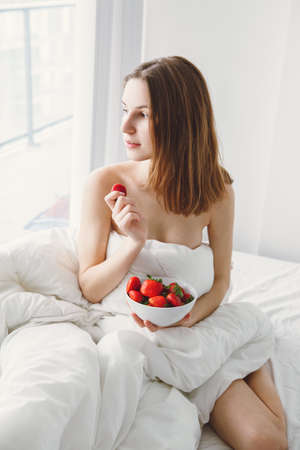 Portrait of smiling beautiful young Caucasian woman with long hair sitting in bed early morning by window, eating red fresh strawberry, lifestyle, toned with filtersの写真素材