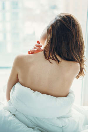 Back view of young Caucasian woman with long hair sitting in bed by window after waking up, indoors...