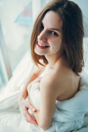 Close up portrait of smiling young beautiful Caucasian woman sitting in bed after waking up, indoors at home early morning, lifestyle, toned with filtersの写真素材