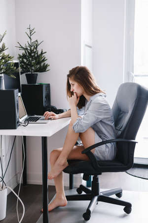 Portrait of young beautiful Caucasian girl woman student in pajamas shirt  working on laptop computer sitting in office chair at home, browsing internet, drinking coffee, early morning lifestyleの写真素材