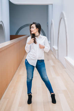 Portrait of smiling white Caucasian brunette young beautiful girl woman model with long dark hair in white shirt and blue jeans indoor in hall of college, university, standing full figure looking awayの写真素材
