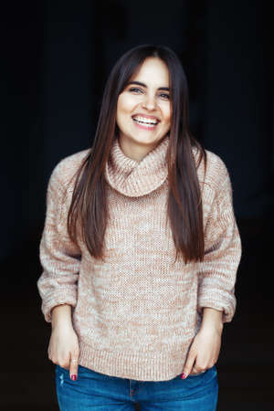 Portrait of smiling laughing white Caucasian brunette young beautiful girl woman model with long dark hair and brown eyes in turtleneck sweater and blue jeans indoor on black backgroundの写真素材