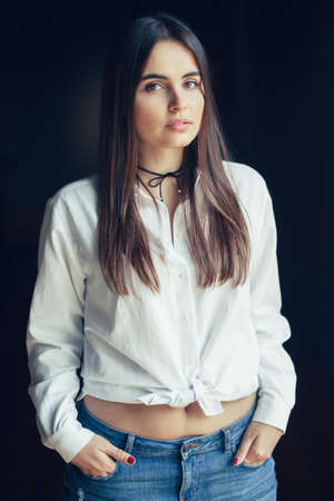 Portrait of pensive sad white Caucasian brunette young beautiful girl woman model with long dark hair and brown eyes in white shirt tied in a knot, blue jeans, looking in camera, on black backgroundの写真素材