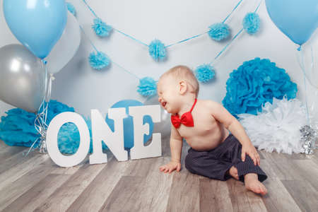 Portrait of crying screaming unhappy Caucasian baby boy in dark pants and red bow tie  on  his first birthday with letters one and balloons, sitting on wooden floor in studioの写真素材
