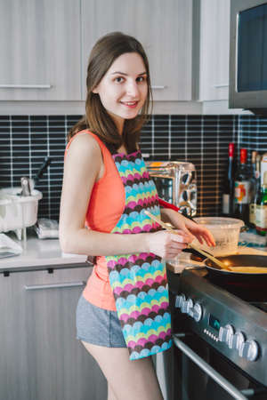 Portrait of young Caucasian woman girl with long hair cooking food pancakes standing in kitchen indoors at home, authentic real people lifestyleの写真素材