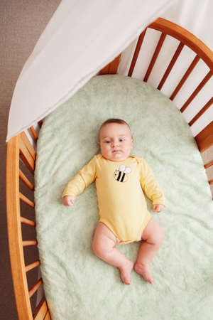 Portrait of cute  funny adorable white Caucasian little baby newborn in yellow shirt lying in crib alone near window, lifestyle candid real life, view from top aboveの写真素材