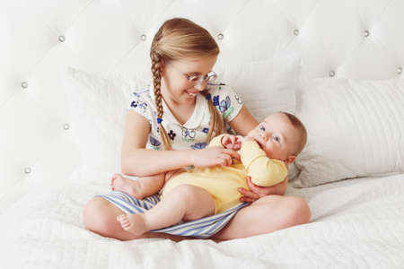Portrait of cute adorable little white Caucasian girl sister with braids playing with her newborn baby brother, sitting on bed in bedroom, lifestyle real true emotions, sibling loveの写真素材