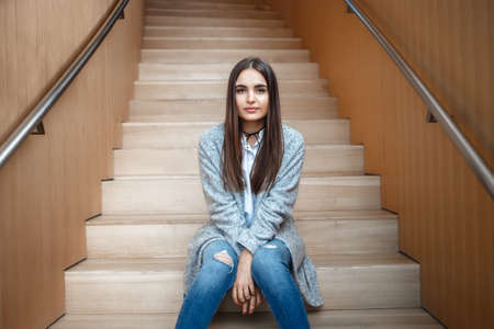 Portrait of pensive Caucasian brunette young beautiful girl woman with long dark hair and brown eyes in white shirt, grey coat and blue jeans sitting on staircase looking in cameraの写真素材