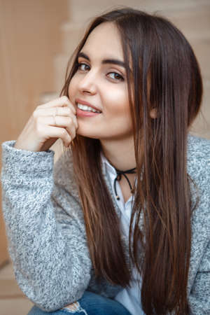 Closeup portrait of smiling Caucasian brunette young beautiful girl woman with long dark hair and brown eyes in white shirt and grey coat sitting on staircase looking in cameraの写真素材