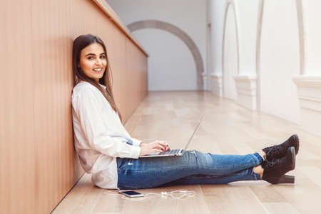 Portrait of  beautiful white Caucasian brunette young girl woman model with long dark hair in white shirt and blue jeans sitting on floor in hall at college university working on laptopの写真素材
