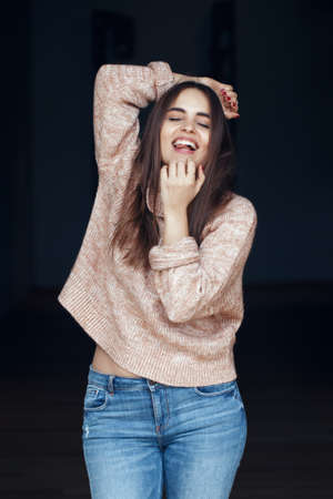 Portrait of smiling laughing white Caucasian brunette young beautiful girl woman model with long dark hair and brown eyes in turtleneck sweater and blue jeans indoor on black backgroundの写真素材