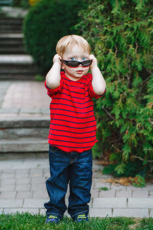 Portrait of funny cute adorable white Caucasian toddler child boy with blond hair and blue eyes in red pullover wearing sunglasses standing in summer park outsideの写真素材