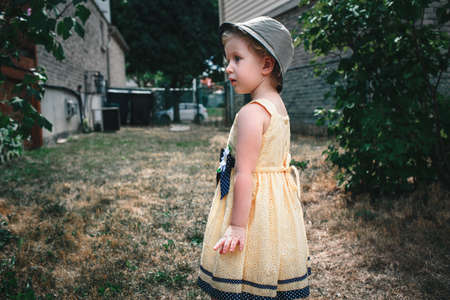 Portrait of cute Caucasian girl wearing yellow dress and hat with funny face expression outside on house backyard on summer day, surprised scared standing among large bushesの写真素材
