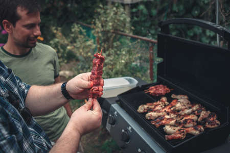 Closeup shot of man person hands holding raw meat skewers ready to fry barbecue, chicken legs thighs on grill fire, party picnic on home backyard, lifestyle tonedの写真素材