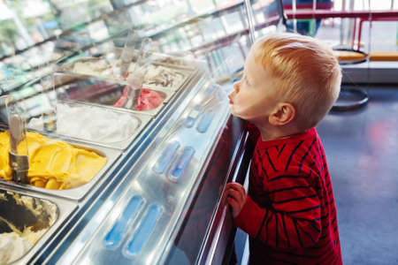 Portrait of cute adorable white Caucasian funny blond child boy looking at ice cream in shop window, trying to choose one, looking surprised puzzled, emotional face expressionの写真素材