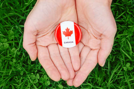 Macro closeup shot of hands palms holding round badge with red white canadian flag maple leaf, on green grass forest nature background outside, Canada Day celebrationの写真素材