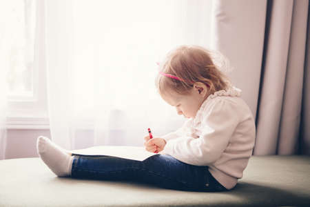Portrait of cute adorable white Caucasian little girl drawing with pencil on paper, sitting on couch...