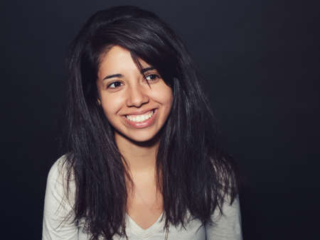 Portrait of beautiful smiling Hispanic latino girl woman with dark brown eyes long dark messy untidy hair in white shirt posing in studio on dark black background, looking awayの写真素材