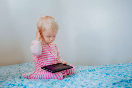 Cute adorable white Caucasian blond baby girl sitting in bed playing with digital tablet with funny face expression, candid lifestyle, early development, new technology generationの写真素材